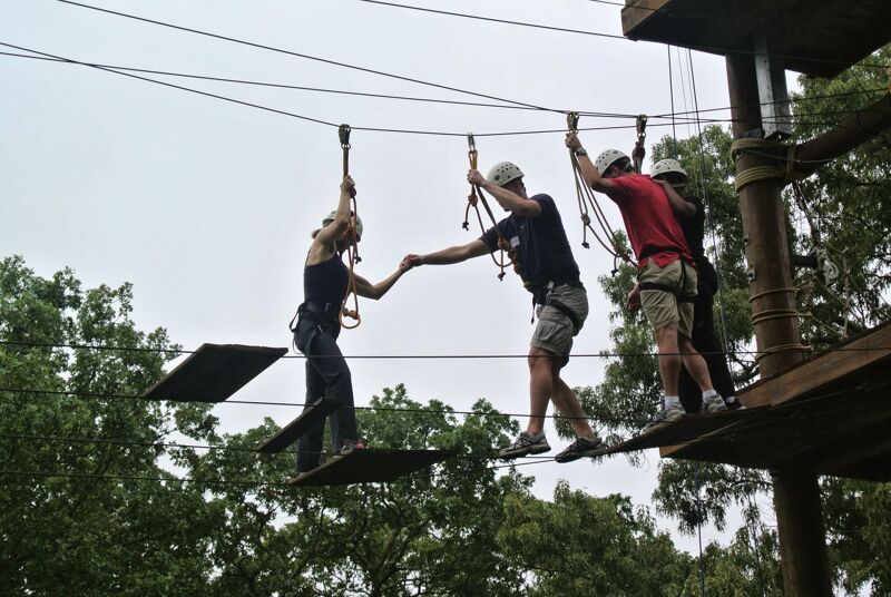 The image shows three people on a ropes course. They are wearing helmets and harnesses, and they are walking across a series of wooden platforms suspended in the air. The course is set in a forest, and the trees provide a natural backdrop. The sky is overcast, and the light is soft.